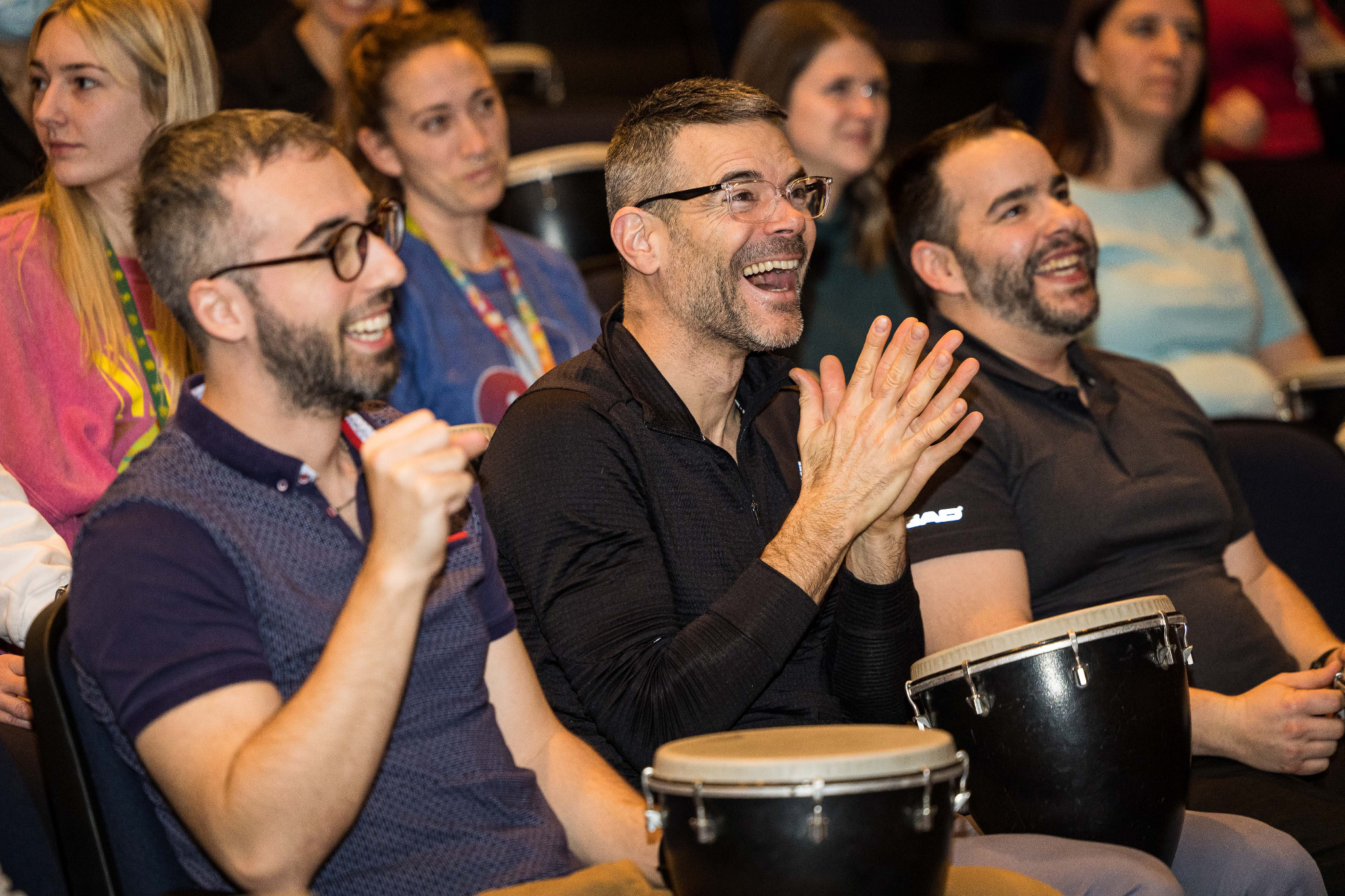 Trois participants souriants avec leurs djembés dans un auditorium. / Three smiling participants with djembes in an auditorium.