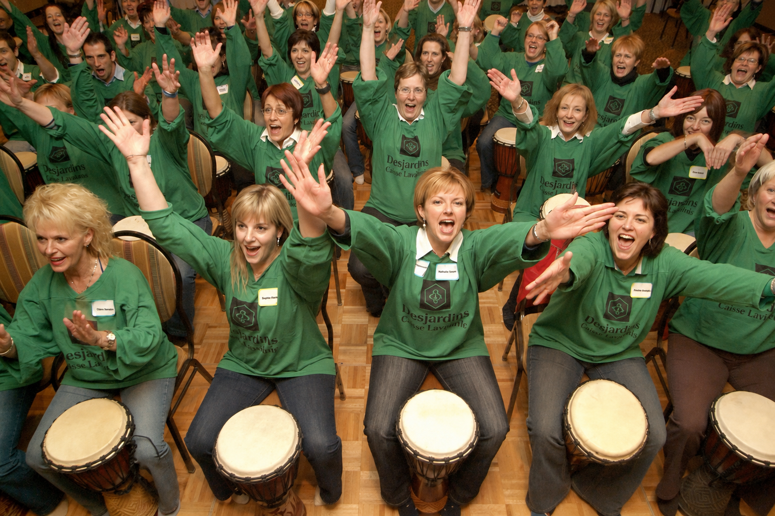 Un groupe en chandails verts lève les bras derrière ses djembés. / A group in green shirts raises their arms behind their djembes.