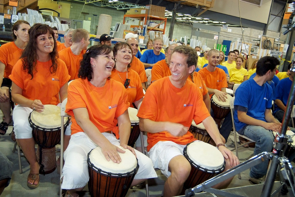 Des participants en chandails orange et bleus jouent du djembé en usine. / Participants in orange and blue shirts play djembes in a factory.