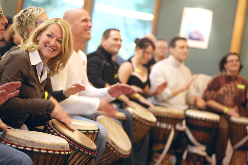 Des collègues souriants jouent des djembés traditionnels dans une salle lumineuse. / Smiling colleagues play traditional djembes in a bright room.