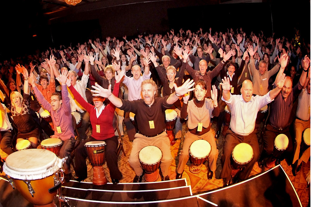 Un grand public de participants au djembé lève les bras dans un auditorium. / A large audience of djembe participants raises their arms in an auditorium.