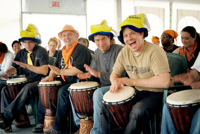 Des participants en chapeaux fantaisie jouent du djembé sous un chapiteau. / Participants wearing festive hats play djembes under a marquee.