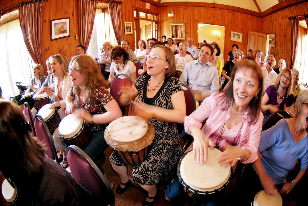 Un groupe souriant joue des djembés dans une salle lambrissée. / A smiling group plays djembes in a wood-panelled room.