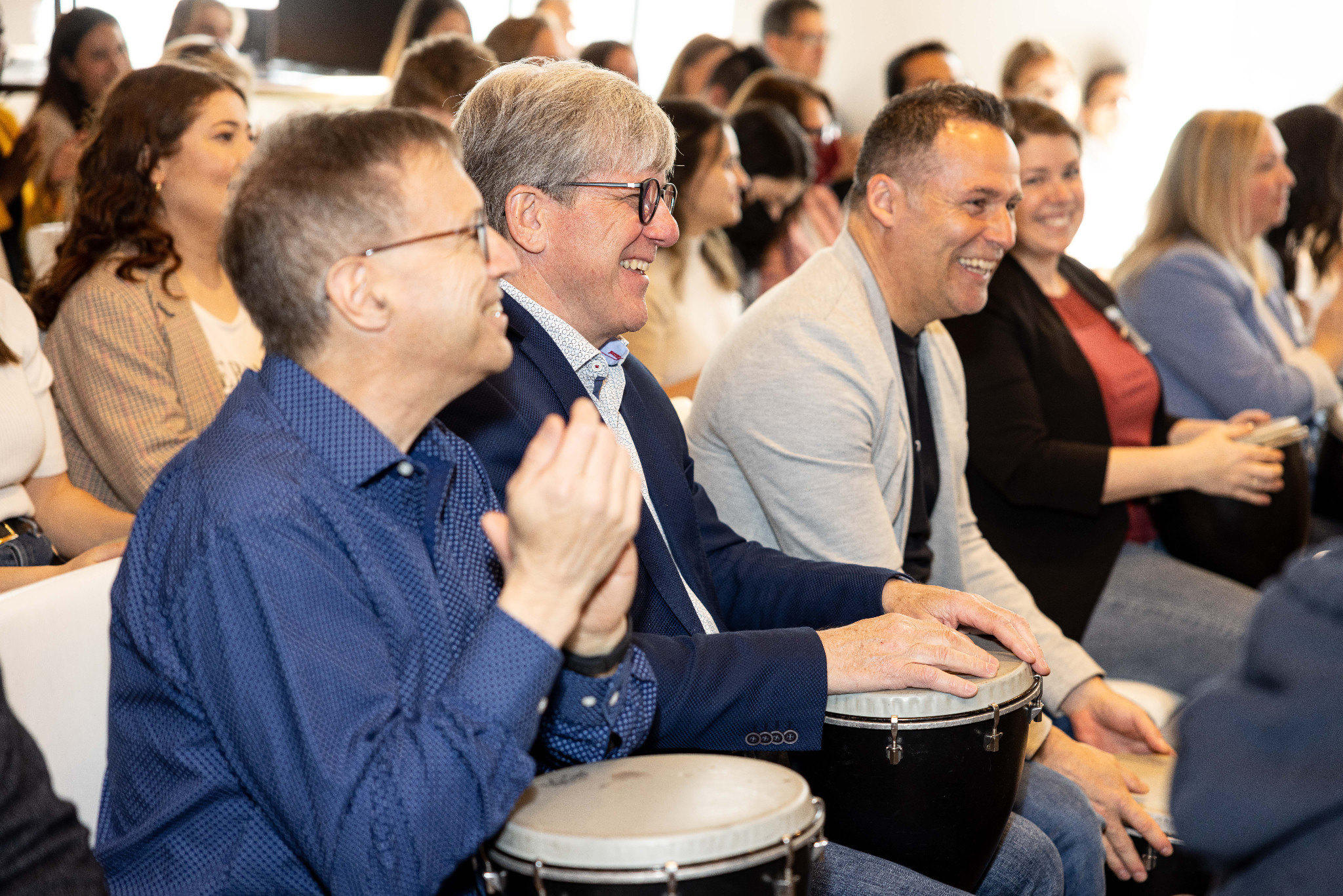 Trois hommes souriants jouent des djembés noirs avec leurs collègues. / Three smiling men play black djembes with their colleagues.